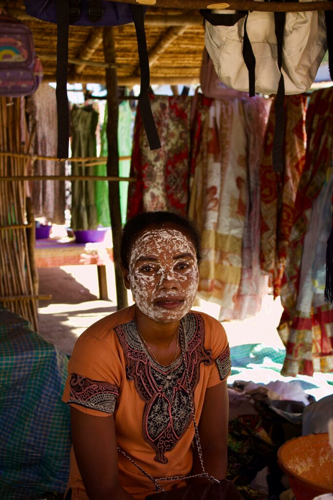pexels photo 34123056 34123056 Woman with a traditional face mask in a Malagasy market stall displaying vibrant fabrics.